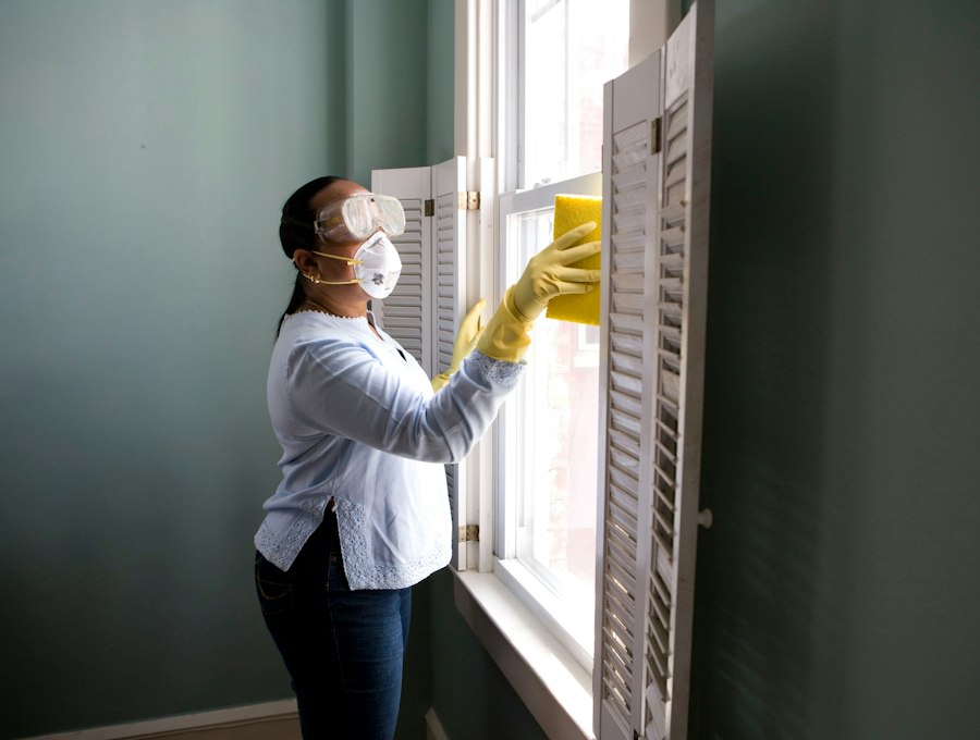 A maid in protective gloves cleaning a window and shutter in a sunlit room